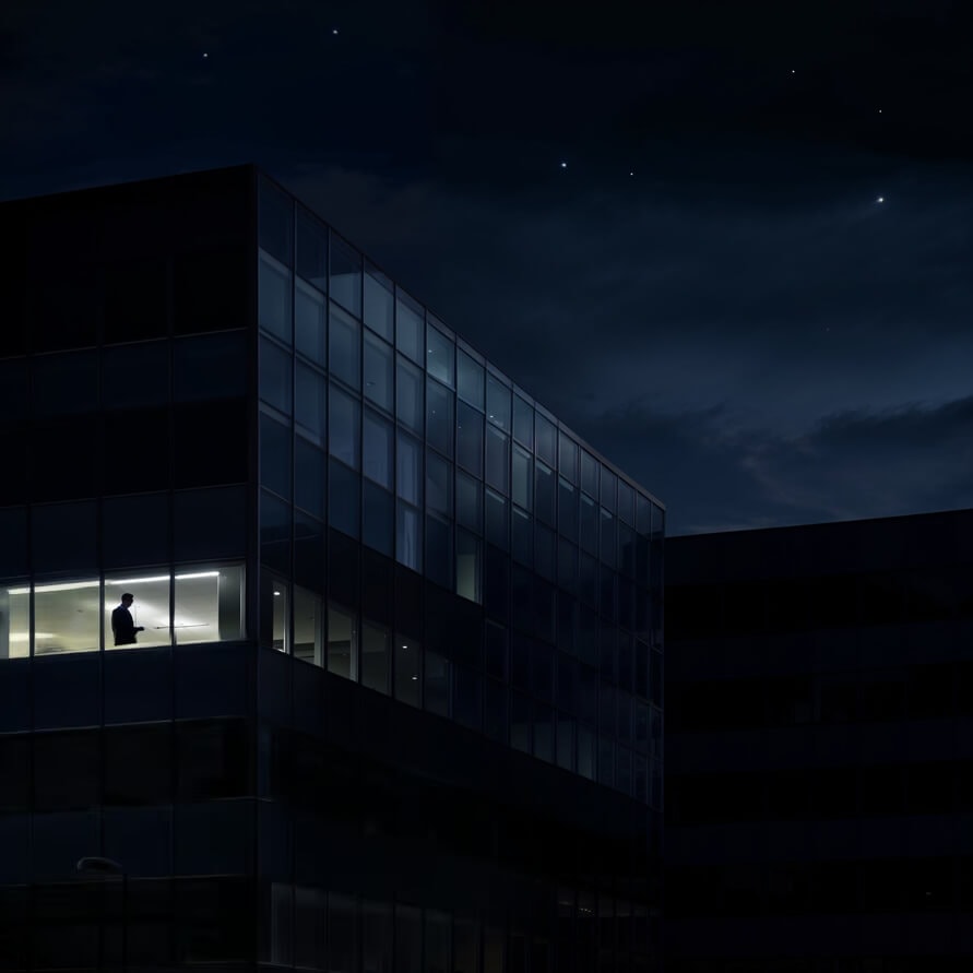Ein Bürogebäude bei Nacht unterm Sternenhimmel. Nur ein Stockwerk ist erleuchtet. Ein nachdenklicher Mann steht am Fenster und denkt über dein Design nach.