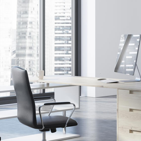 Ein helles Büro mit Drehstuhl, Schreibtisch, weisser Kaffeetasse und einem 24“iMac, in dessen Screen sich die Fensterfronten anderer Bürogebäude spiegeln.