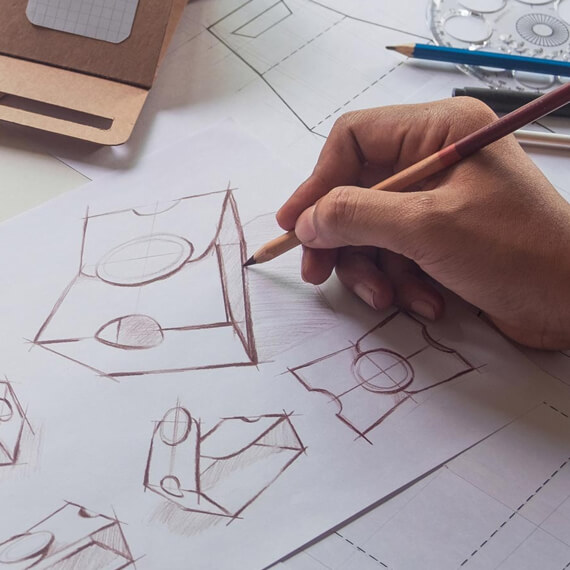 Eine jüngere männliche Hand führt einen Bleistift über das Papier und skizziert ein aufgeklapptes Verpackungsdesign in unterschiedlichen Perspektiven.