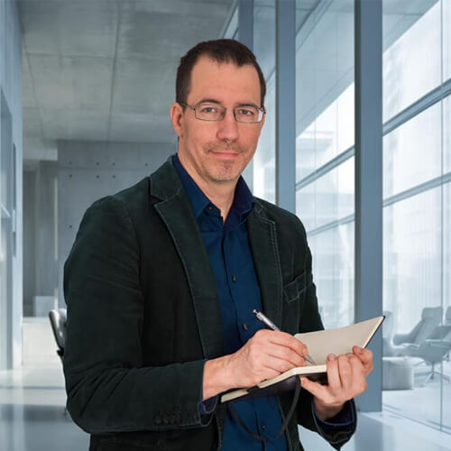 Stephan Bender mit Brille und schwarzem Jacket. In seiner Hand hält er ein Notizbuch und einen Stift. Der Grafiker steht hier in einem modernen Büroflur.